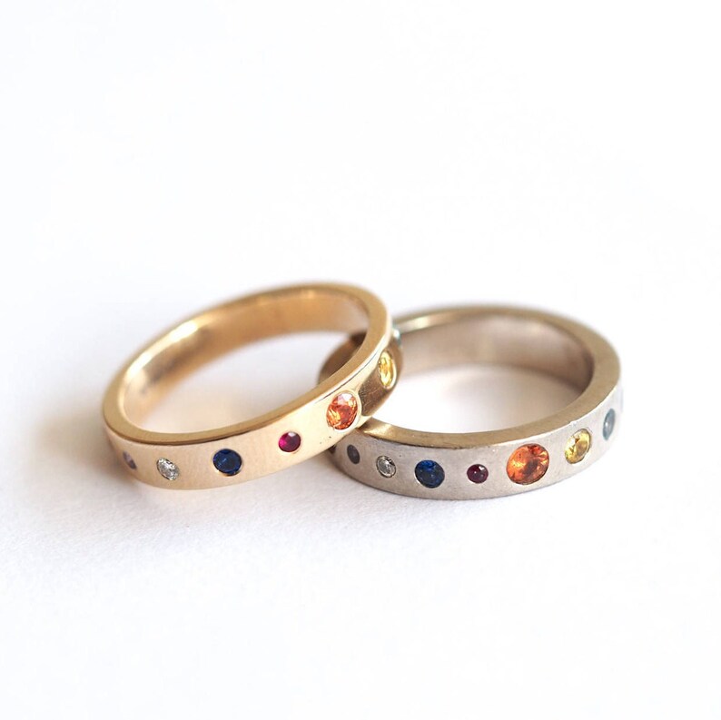 May include: Two rings with colorful gemstones. One ring is gold-toned, the other is silver-toned. Both rings have a series of small, round gemstones in various colors, including red, blue, yellow, and orange. The rings are set against a white background.