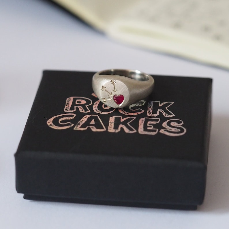Puede incluir: Un anillo de sello de plata con un coraz&oacute;n rojo y flechas grabadas en la parte superior. El anillo est&aacute; sentado en una caja negra con las palabras "Rock Cakes" impresas en la parte superior.