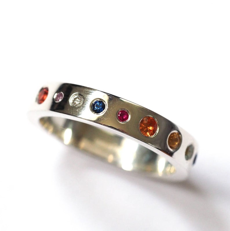 May include: A silver ring with a series of colorful gemstones. The gemstones are round and include red, orange, yellow, blue, and pink. The ring is set against a white background.