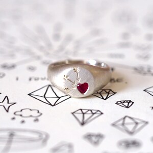 Puede incluir: Un anillo de sello de plata con un coraz&oacute;n rojo y flechas grabadas en la parte superior. El anillo est&aacute; sobre un fondo blanco con dibujos negros de diamantes.