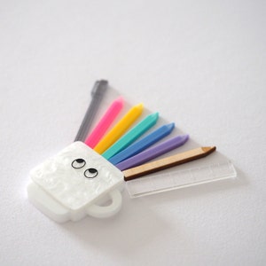 May include: A white ceramic mug with a face and two black eyes, with a rainbow of colored pencils and a ruler sticking out of the top.