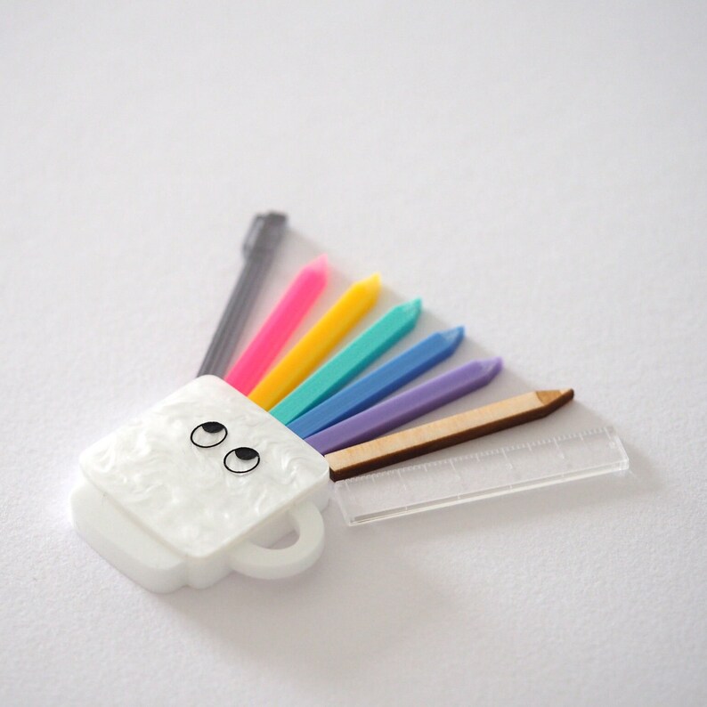 May include: A white ceramic mug with a face and two black eyes, with a rainbow of colored pencils and a ruler sticking out of the top.