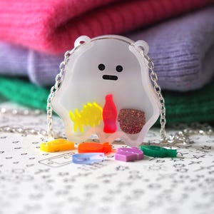 Puede incluir: Un collar de resina transparente con un oso blanco con la cara negra y un alimento rojo, amarillo y marrón en su interior. El collar está en una cadena plateada y hay cinco pequeños amuletos de resina de colores en la mesa.