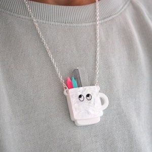 May include: A silver chain necklace with a white ceramic mug pendant. The mug has a face with two black eyes and a black outline. Inside the mug are three colorful pencils.