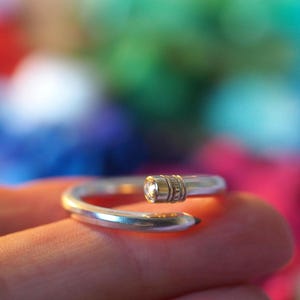 May include: A silver ring in the shape of a pencil, with a clear gemstone at the top and a pencil tip at the other end. The ring is displayed on a finger, set against a blurred, colourful background.