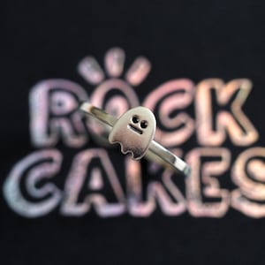 Może przedstawiać: Srebrny pierścionek z zawieszką w kształcie ducha. Duch ma dwa małe czarne oczy i prosty uśmiech.