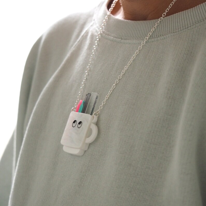 May include: A white ceramic mug necklace with a silver chain. The mug has two black eyes and is filled with colorful pencils.