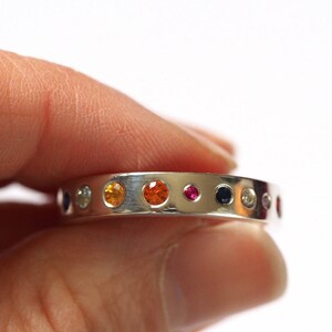 May include: A silver ring with a series of colorful gemstones. The gemstones are in various colors, including orange, yellow, blue, and pink. The ring is held by a person's fingers.