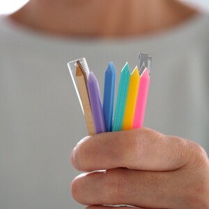 May include: A hand holding a variety of colorful pencils and a wooden ruler. The pencils are blue, green, pink, and yellow. The ruler is made of wood and has a metal tip.