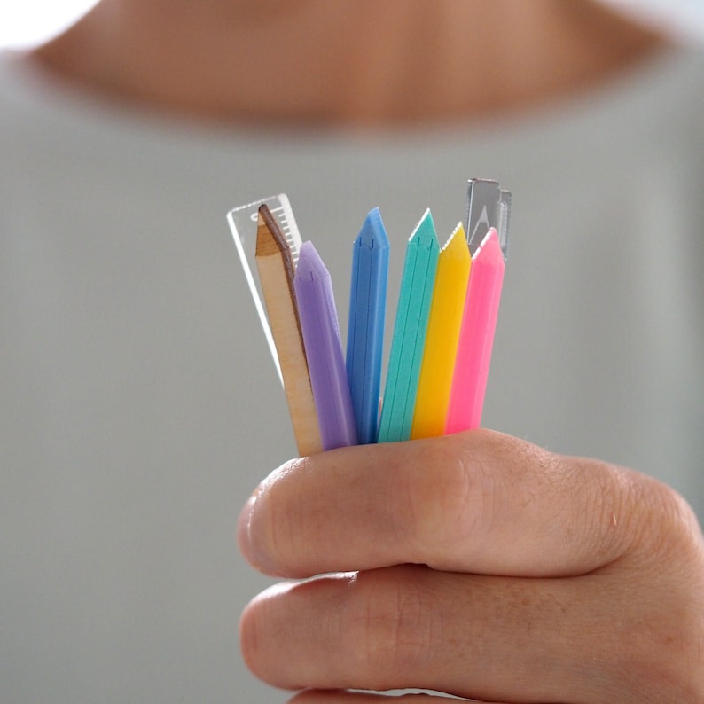 May include: A hand holding a variety of colorful pencils and a wooden ruler. The pencils are blue, green, pink, and yellow. The ruler is made of wood and has a metal tip.