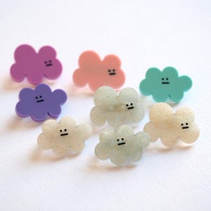 May include: A selection of cloud-shaped accessories in shades of purple, pink, and turquoise. Each cloud features a simple black face. Some have a glittery finish, adding a touch of sparkle.