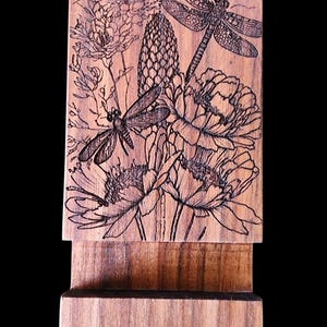 Puede incluir: Soporte de teléfono de madera con un grabado detallado de libélulas y flores. El diseño está grabado en la madera, creando un contraste. El soporte está hecho de madera de tonos cálidos y tiene un diseño simple y funcional.