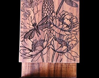 Puede incluir: Soporte de teléfono de madera con un grabado detallado de libélulas y flores. El diseño está grabado en la madera, creando un contraste. El soporte está hecho de madera de tonos cálidos y tiene un diseño simple y funcional.