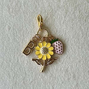 Peut inclure: Un charm doré avec un tournesol jaune, une fraise rose et la lettre "B". Le tournesol a un centre marron. La fraise a des feuilles vertes. Le charm est attaché à un fermoir doré, idéal pour un bracelet ou un collier.