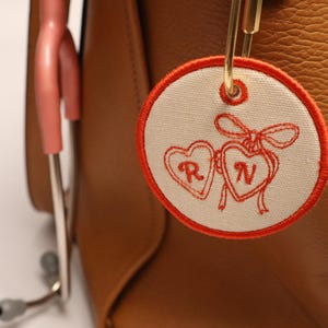 Peut inclure: Un porte-clés rond en tissu blanc cassé avec une bordure brodée rouge et deux cœurs, chacun contenant une lettre. L'étiquette est attachée à un fermoir doré sur un sac en cuir marron. Un stéthoscope rose est visible en arrière-plan.