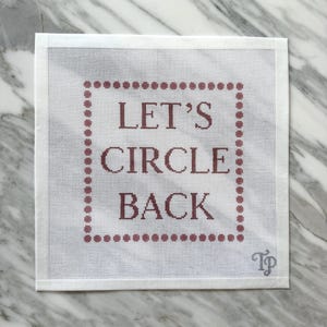 May include: A square white needlepoint canvas featuring the text LET'S CIRCLE BACK in dark red letters. The text is enclosed within a square border made of small dark red dots. A small grey TP logo is visible in the bottom right corner of the canvas.