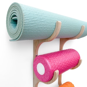 May include: A wooden wall-mounted rack with three hooks for storing yoga mats and foam rollers. The top hook holds a rolled up light blue yoga mat. The middle hook holds a pink foam roller. The bottom hook is empty.