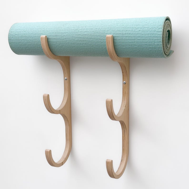 May include: A wooden wall-mounted yoga mat rack with two hooks. The rack is made of light-colored wood and has a natural finish. The hooks are designed to hold yoga mats securely in place.