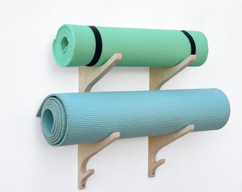 May include: Two rolled yoga mats, one mint green and one light blue, mounted on a wooden wall rack. The green mat has a black strap. The rack is made of light-colored wood and is attached to a white wall.