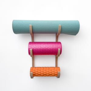 May include: A wooden wall-mounted rack with three shelves holding a light blue yoga mat, a pink foam roller, and an orange foam roller.