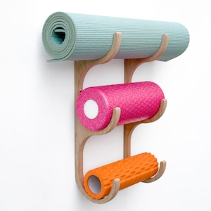 May include: A wooden wall-mounted rack with three hooks for storing yoga mats and foam rollers. The rack has a light wood finish and is holding a light blue yoga mat, a pink foam roller, and an orange foam roller.