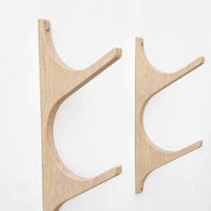 May include: Two wooden wall-mounted surfboard racks. The racks are made of light-colored wood and have a curved design. They are mounted on a white wall.