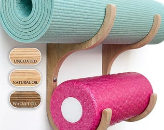 May include: A wall-mounted wooden rack with two curved tiers holding a rolled light blue yoga mat on top and a textured bright pink foam roller on the bottom. Three oval labels show wood finish options labelled Uncoated, Natural Oil, and Walnut Oil.