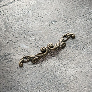 以下が含まれることがあります： アンティーク真鍮色の小さな装飾用金属要素。対称的なデザインで、渦巻き模様と葉のようなディテールが特徴で、クラフトプロジェクトや家具の装飾に適しています。