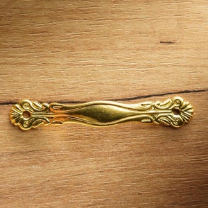 May include: A gold-colored metal decorative accent with a detailed ornate design. The accent has a long, narrow shape with a slightly curved center.