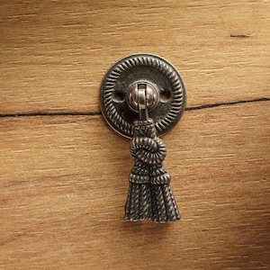 Puede incluir: Un tirador de cajón de metal plateado con un borla decorativa. El tirador está montado en una superficie de madera.