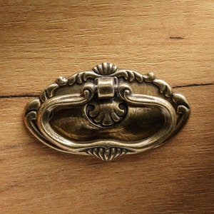 Puede incluir: Un tirador de cajón de latón de estilo vintage con un diseño ornamentado. El tirador tiene forma ovalada con un centro elevado y bordes decorativos.