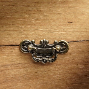 Può includere: Una maniglia per cassetto in metallo decorativa con un design a scorrimento e una finitura nera. La maniglia ha due accenti dorati su ogni lato.