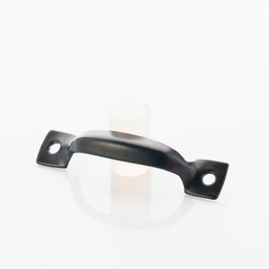 Puede incluir: Pomo de metal negro con bordes redondeados y dos agujeros en cada extremo. El pomo está sobre una superficie blanca.