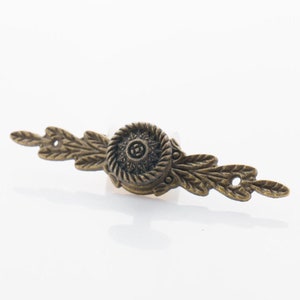 Può includere: Una maniglia per cassetto decorativa in bronzo con un design floreale. La maniglia ha un pomo rotondo centrale con un motivo dettagliato e due estensioni a forma di foglia su entrambi i lati.