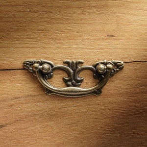 Może przedstawiać: Stylizowany, ozdobny, mosiężny uchwyt do szuflady w stylu vintage. Uchwyt jest zamontowany na drewnianej powierzchni.