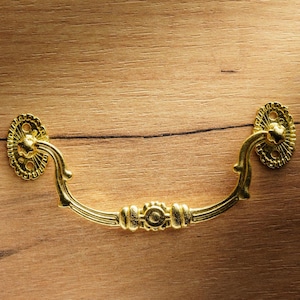 Può includere: Una maniglia per cassetto dorata con un design decorativo. La maniglia è fissata a una superficie in legno.