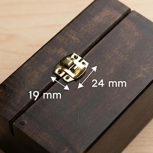 Puede incluir: Una caja de madera teñida oscura con un cierre de color latón. La caja es rectangular y mide 19 mm por 24 mm. La caja está cerrada y se encuentra sobre una superficie de madera clara.