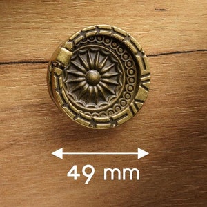 Op de afbeelding: Een ronde, sierlijke, messingkleurige kastknop met een decoratief bloemmotief. De knop heeft een diameter van 49 mm.
