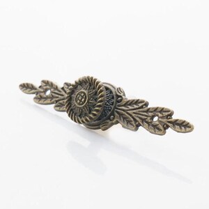 May include: A decorative bronze drawer pull with a floral design. The pull has a central circular element with a raised design and two leaf-like extensions on either side.