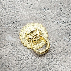 以下が含まれることがあります： ライオンの頭部の装飾が施された金色のドアノブ。ノブは灰色のテクスチャのある表面にあります。