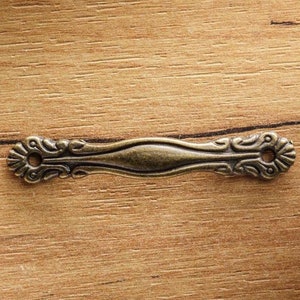 May include: A decorative metal pull with an ornate design in a bronze finish. The pull has two holes for attaching to a drawer or cabinet.