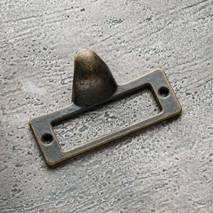 Puede incluir: Un portaetiquetas de latón con una parte superior puntiaguda y una abertura rectangular para una etiqueta. El portaetiquetas está sobre una superficie gris texturizada.