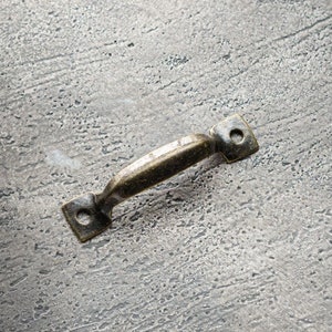Puede incluir: Un pequeño tirador de metal de estilo antiguo con un agarre curvo y placas de montaje cuadradas. El tirador tiene un acabado bronce y está colocado sobre una superficie gris texturizada, lo que sugiere una estética vintage o rústica.
