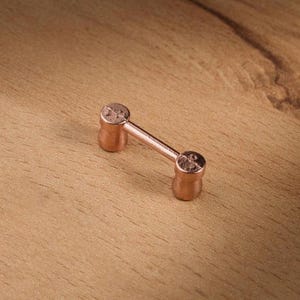 Può includere: Un piccolo oggetto a forma di manubrio color rame, con due estremità arrotondate collegate da una barra dritta. L'oggetto è appoggiato su una superficie di legno marrone chiaro, con la venatura del legno visibile.