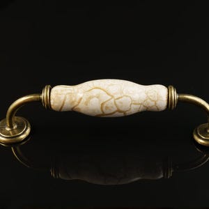 May include: A cabinet pull featuring a cream-coloured, crackled ceramic handle and antique brass hardware. The handle is curved and has a decorative, textured surface. The pull is designed for furniture and cabinetry.