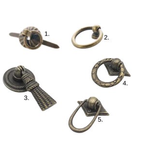 Op de afbeelding: Vijf antieke bronzen kasttrekkers in verschillende ontwerpen. De trekkers omvatten een ringtrekker, een druppelvormige trekker en trekkers met decoratieve achterplaten. De hardware is ideaal om een vintage touch aan meubels toe te voegen.