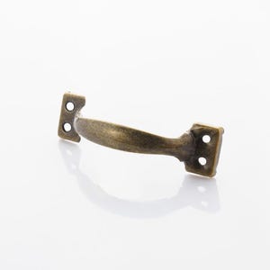 Puede incluir: Un tirador de armario de color bronce con un asa curvada y dos placas de montaje, cada una con dos agujeros. El herraje tiene una estética vintage y está diseñado para muebles o armarios.