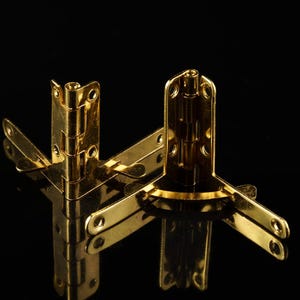Könnte beinhalten: Drei goldfarbene Eckbeschläge vor schwarzem Hintergrund. Die Scharniere sind so konzipiert, dass sie zwei Holzstücke in einem 90-Grad-Winkel verbinden.