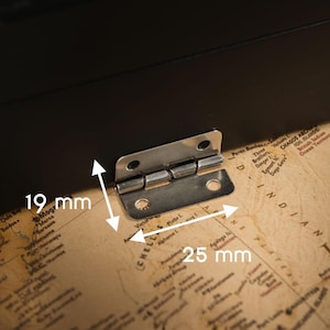 May include: A silver-colored metal hinge, 25 mm wide and 19 mm high, is attached to a black surface. The hinge is mounted on a map-printed surface. The hinge has a rectangular shape with rounded corners and multiple screw holes.