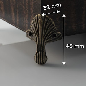 May include: Close-up of a decorative, antique-style metal furniture foot. The foot is bronze-colored, with a leaf-like design. The foot measures 45 mm in height and 32 mm in width. The wooden furniture is dark brown.